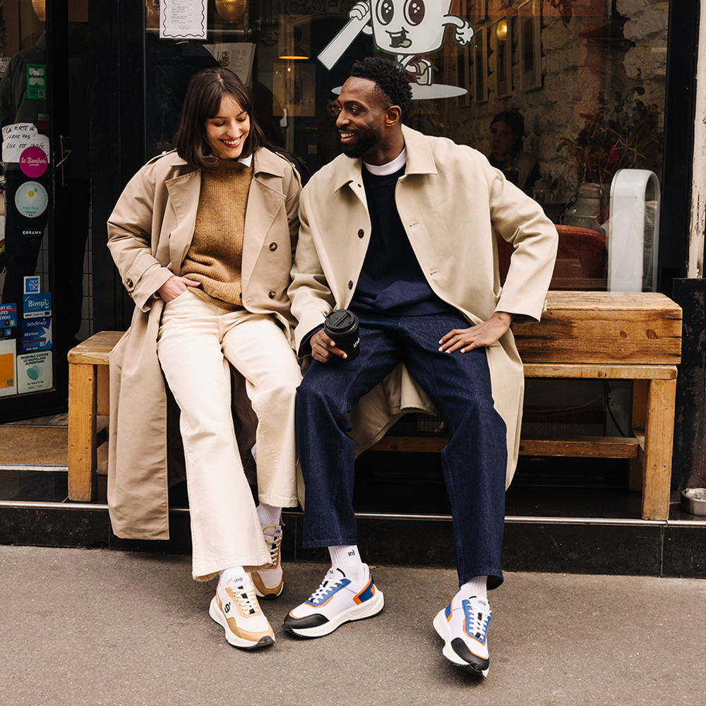 Un couple assis côte à côte souri. Ils se situent devant un café parisien, gobelet de café en main.