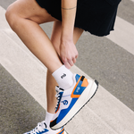 Femme portant des sneakers orange, ajustant sa chaussette "iné" de la marque iné, disponibles du 36 au 46